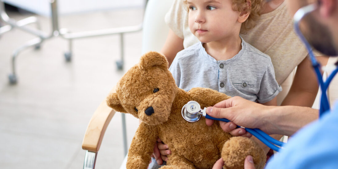 Little Boy Visiting Doctor Portrait of adorable curly child in doctors office holding teddy bear toy, with pediatrician listening to heartbeat using stethoscope